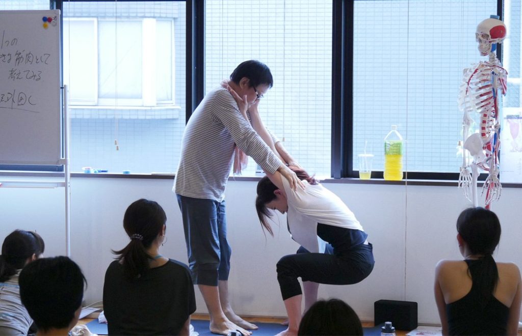 ヨガ解剖学テーマ別肩・股関節の連動の講座風景。講師(内田かつのり)が中腰の生徒が向かい合って立ち7、講師の肩に両腕をかけてぶら下がるように腰を落としている様子